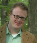 A man with short reddish-blonde hair and glasses smiling while wearing a light green shirt and tan jacket.