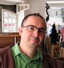 A man with short dark hair and glasses wearing a green shirt and brown jacket.