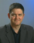 A man with short dark hair wearing a dark blazer and a dark collared shirt against a blue background.