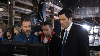 Three men in a warehouse look intently at a screen in a dimly lit industrial space.