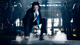 A man in a dark suit and blue tie crouches on a platform amidst smoke in a dimly lit warehouse.