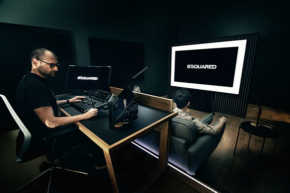 A man sits at a desk with multiple monitors and a control panel, facing a large screen in a dark room.