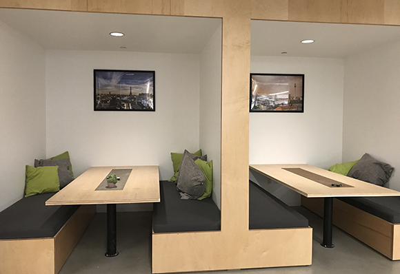 Two wooden office pods containing light wood tables, dark gray benches, and green and gray pillows.