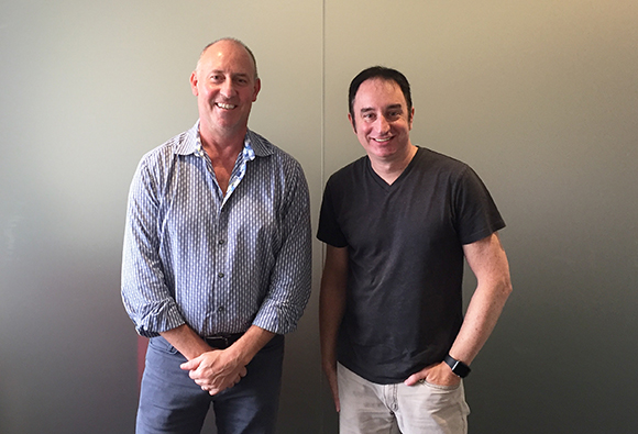 Two men stand smiling against a gray wall. The man on the left wears a striped button-down shirt and the man on the right wea