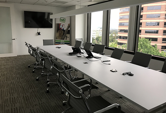 A long white conference table surrounded by black office chairs in a bright room with large windows.