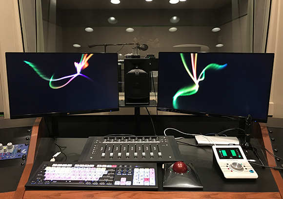 Dual computer monitors, a mixing console, and a control surface on a desk in a recording studio.