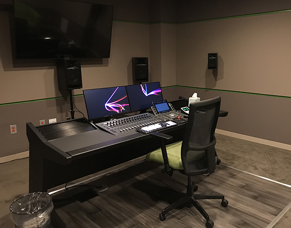 A black mixing console with dual monitors, studio monitors, and a mesh office chair in a dim studio.