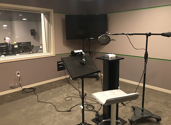 A recording studio room with a microphone on a stand, a black pedestal, a stool, and a large monitor.