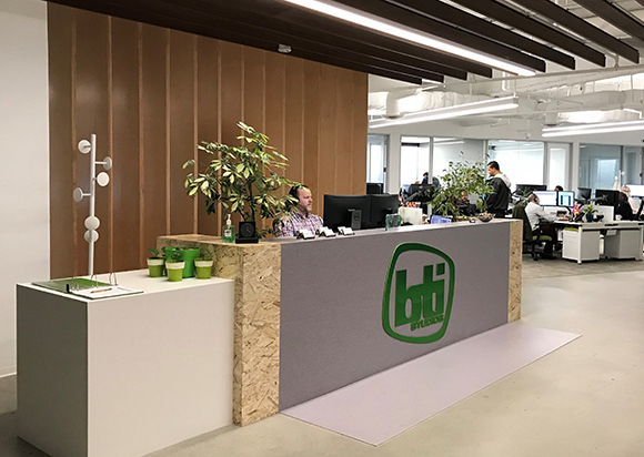 A modern office reception desk with a gray front featuring a green BTI Studios logo and a wooden accent side.