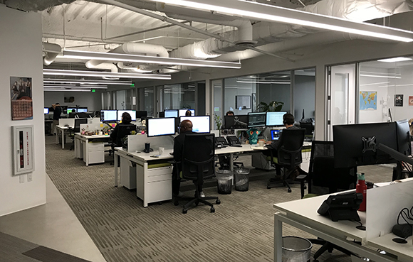 People working at desks with computer monitors in a modern open-plan office with industrial ceilings.