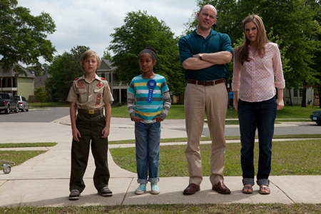 A boy in a scout uniform, a girl with a blue ribbon, a man with crossed arms, and a woman standing on a sidewalk.