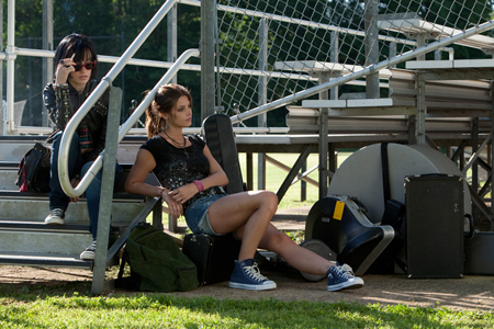 Two young women sit on metal bleachers near a chain-link fence with various bags nearby.