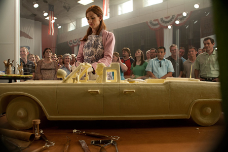 A woman in a floral apron decorating a large, tan car-shaped cake in front of a crowd.