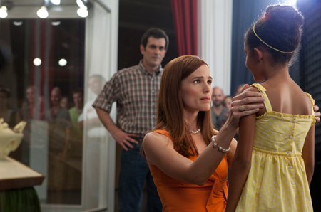 A woman in orange looks intently at a young girl in a yellow dress, with a man standing behind them.