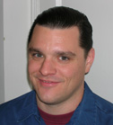A man with dark hair slicked back, smiling, wearing a blue collared shirt over a maroon undershirt.
