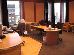 An office interior with wooden desks, black office chairs, and large windows overlooking city buildings.