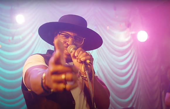 A man wearing a black hat and glasses sings into a microphone while pointing toward the camera.