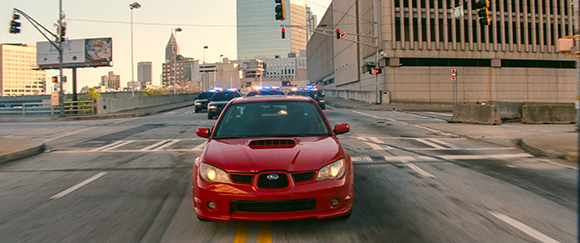 A red Subaru drives toward the camera on a city street with police cars following in the background.