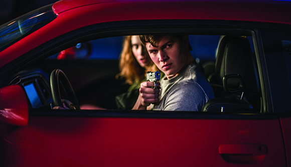 A man in a red car points a handgun toward the camera while a woman sits beside him.