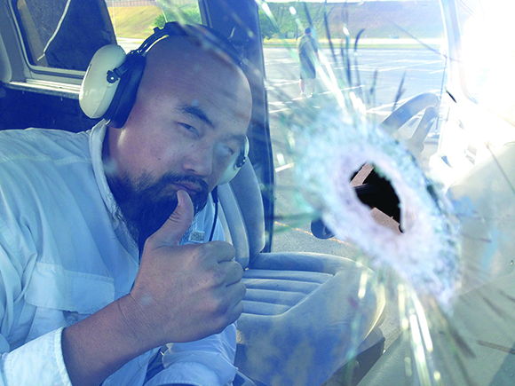 A man with a beard and headphones gives a thumbs up through a windshield with a large bullet hole.