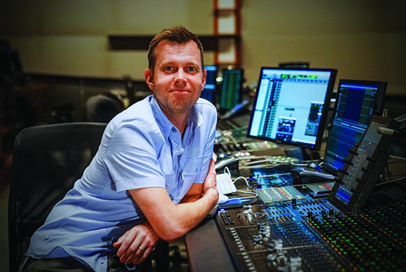 A man in a light blue shirt leans on a large audio mixing desk in a studio with multiple computer monitors.