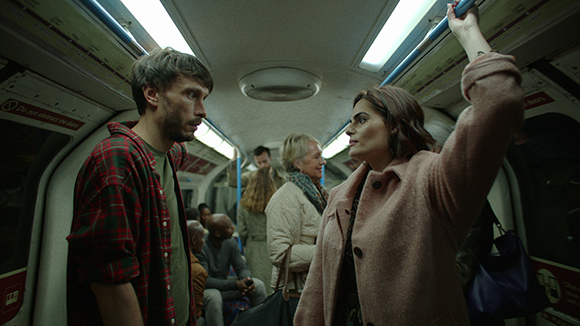 A man in a red plaid shirt and a woman in a pink coat stand facing each other on a crowded subway train.