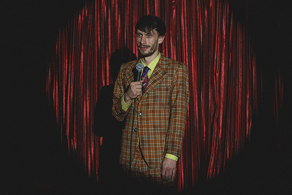 A man in an orange plaid suit holds a microphone against a backdrop of red velvet curtains.
