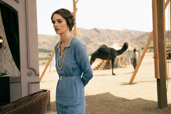 A woman in a blue dress stands outdoors near a tent and a camel in a desert landscape.