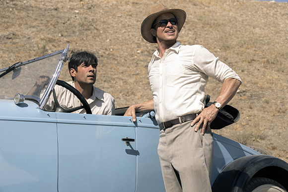A man in a sun hat and sunglasses stands by a light blue vintage car while another man sits in the driver's seat.