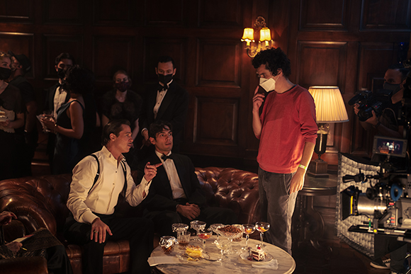 A man in a red shirt stands near men seated on a leather sofa in a dark, wood-paneled room.