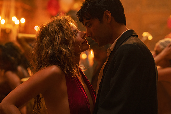 A man in a dark suit and a woman with curly hair lean toward each other in warm, dim lighting.