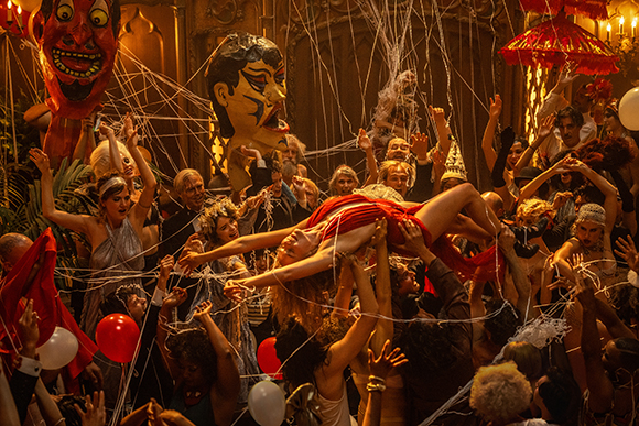 A group of people in costumes dancing and celebrating in a room filled with spiderwebs and large masks.