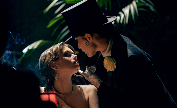 A man in a black top hat and white gloves leans toward a woman in a dark, tropical-themed setting.