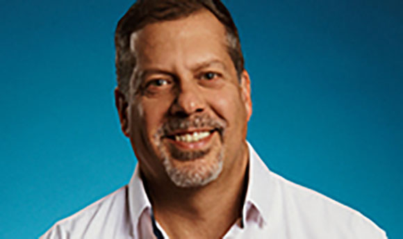 A smiling man with short brown hair and a goatee wearing a white collared shirt.