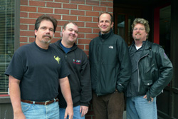 Four men standing together outdoors in front of a red brick wall.