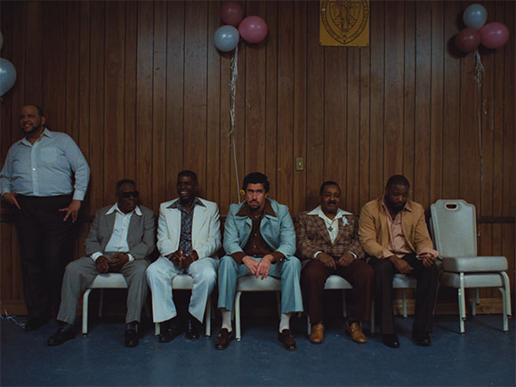 Six men in suits and dress shirts pose in a row against a wood-paneled wall with pink and blue balloons.