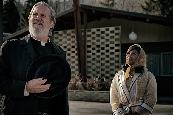 A man in a black priest's collar holds a hat while a woman in a yellow headscarf looks up beside him.