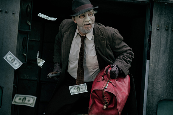 A man in a fedora and brown coat holds a red bag amidst falling dollar bills.