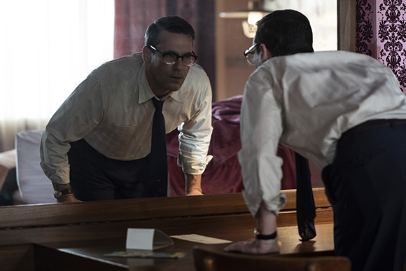 Two men in white dress shirts and dark trousers lean over a wooden table in a dimly lit room.