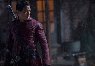 A man in a red leather jacket with a sword on his back stands in the rain at night.