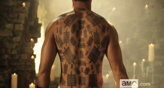 A shirtless man stands with his back to the camera, displaying large black geometric tattoos across his back.