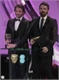 Two men in black suits and ties standing on a stage holding gold awards.