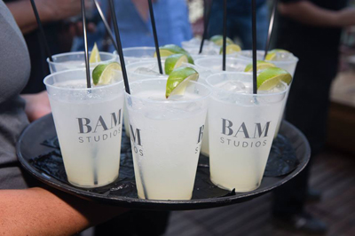 A person holds a black tray filled with several clear plastic cups of white liquid, each with a lime wedge and black straw.