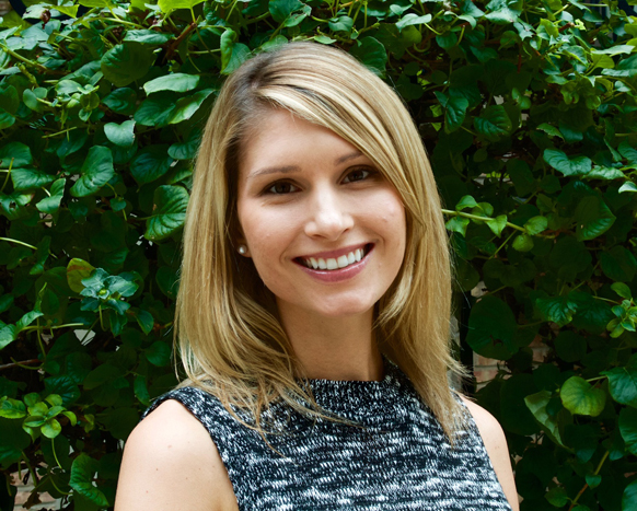 A smiling woman with blonde hair wearing a black and white patterned top, standing in front of green leaves.