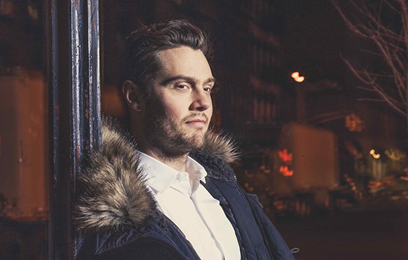 A man with short dark hair and a beard wearing a white shirt and a dark jacket with a fur collar at night.