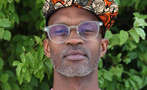 A man with glasses and a patterned cap looking forward against a background of green leaves.