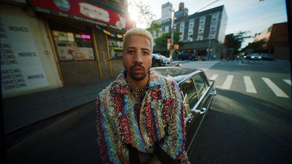 A man with blonde hair wearing a multicolored textured jacket stands on a city street near a crosswalk.