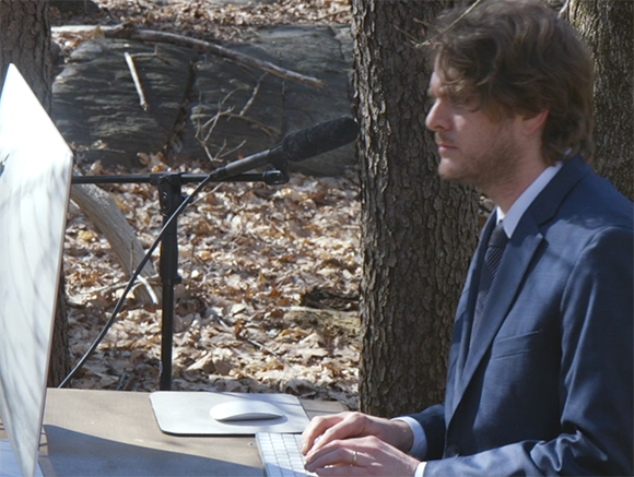 A man in a blue suit sits at a table using a laptop and mouse in a wooded area.