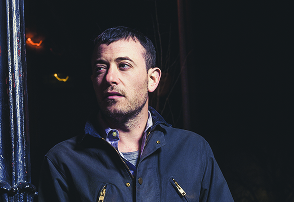 A man with short dark hair and stubble wearing a dark jacket, looking to his right against a dark background.