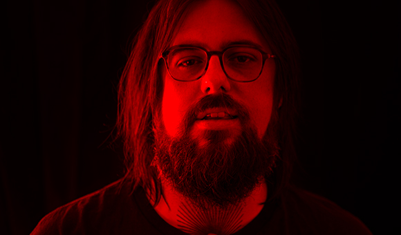 A man with long hair, glasses, and a full beard lit by intense red light against a black background.